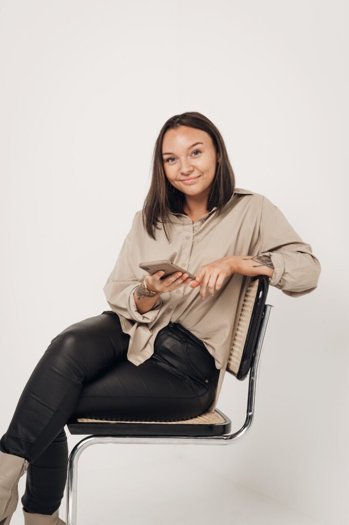 Studiofotografering frendberg akademi med Frendberg Agency blogg. Mathilda sitter på en stol och håller i en telefon och har svart och beiga kläder.